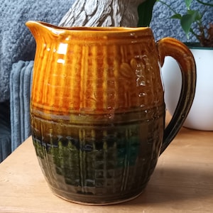 May include: A brown and green ceramic pitcher with a textured surface. The pitcher has a wide mouth and a handle.