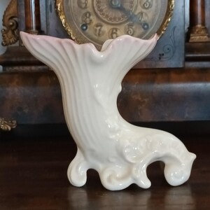 May include: A white ceramic vase with an unusual, organic form. The vase has a flared top with a soft pink tint and a textured, ribbed body. The base features a stylized, curved design. The vase is displayed on a dark wooden surface.