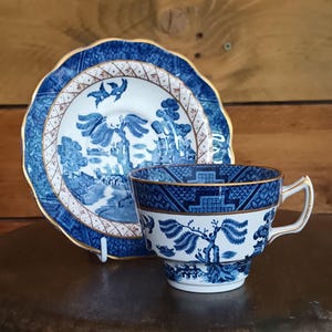 Op de afbeelding: Een blauw-wit porseleinen theekop en schotel set. De kop en schotel hebben een gedetailleerd landschapsontwerp met een gouden rand. De schotel heeft een geschulpte rand. De set wordt gepresenteerd op een donkerbruin oppervlak.