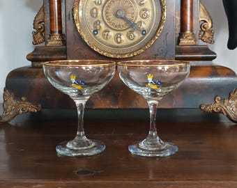 May include: Two clear glass coupe glasses with gold-toned rims. Each glass features a small, colorful graphic of a leaping deer with yellow and blue details on the bowl. The glasses have a short stem and a circular base, designed for serving drinks.