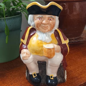 Pode incluir: Uma jarra de cerâmica em forma de homem usando um chapéu de três pontas, um colete amarelo e uma jaqueta marrom. Ele está segurando uma caneca na mão direita e sentado em um toco de árvore.
