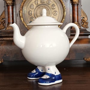 Könnte beinhalten: Eine weiße Keramik-Teekanne mit kugelförmigem Körper, gebogenem Ausguss, Henkel und Deckel. Die Teekanne hat zwei Beine an der Basis, die jeweils einen blauen Schuh mit weißen Akzenten tragen. Das Design umfasst einen kleinen Knopf am Deckel.