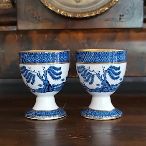 Op de afbeelding: Twee bijpassende eierdopjes. Elk dopje heeft een witte basis en een blauw-wit patroon met een boommotief. De randen zijn goudkleurig afgewerkt. De eierdopjes staan op een houten oppervlak.
