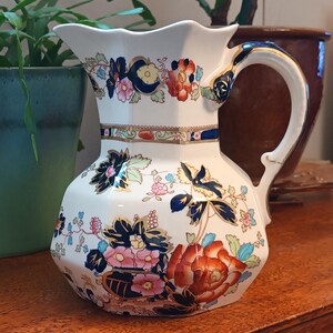 May include: A white ceramic pitcher with a floral design in shades of red, blue, and pink. The pitcher has a curved handle and a scalloped rim. The floral pattern is detailed and covers the body of the pitcher.