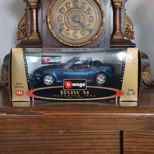 Op de afbeelding: Een blauwe BMW M Roadster 1996 modelauto in een displaydoos. Het model is schaal 1/24. De doos heeft het Burago-logo en de tekst "Bijoux Collection". De auto wordt tentoongesteld voor een vintage klok.