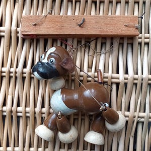 Puede incluir: Títere de perro de madera vintage, pintado en marrón y blanco. El títere tiene una nariz negra y está suspendido por cuerdas de una barra de madera. El fondo es un patrón de mimbre tejido.
