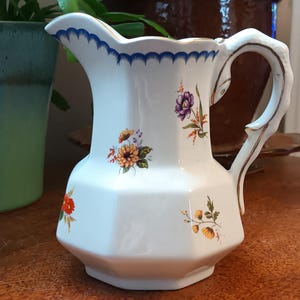May include: White ceramic pitcher with a gold rim and blue and white trim. The pitcher has a floral design with yellow, orange, purple, and green flowers.