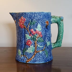 Könnte beinhalten: Eine blaue Keramikkanne mit einem grünen Henkel und einem Blumendesign. Die Kanne hat eine strukturierte Oberfläche und ist mit rosa und grünen Blumen verziert.