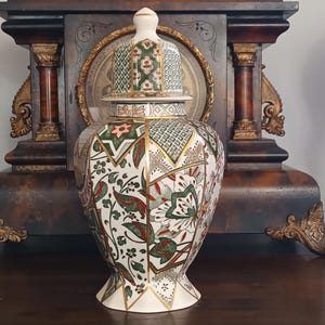 May include: A ceramic ginger jar with a lid, featuring a white base decorated with intricate geometric patterns and floral motifs in shades of green, red, and gold. The jar has a tapered neck, a flared base, and gold-colored trim accents.