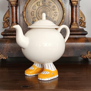 May include: A whimsical, off-white ceramic teapot with a round body, a lid, and a spout. The teapot is supported by a pair of yellow shoes with blue accents. The teapot is displayed on a dark wooden surface.
