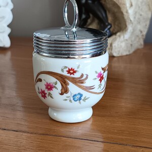Może przedstawiać: Biały ceramiczny pojemnik na jajka z srebrną pokrywką i kwiatowym wzorem. Pokrywka ma uchwyt w kształcie pierścienia.