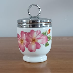 Puede incluir: Un huevero de porcelana blanca con un diseño floral con flores rosas y hojas verdes. El huevero tiene una tapa de metal plateado con un asa de anillo. La base es un pedestal redondeado.