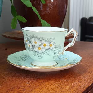 May include: A vintage teacup and saucer set with a delicate floral pattern. The cup and saucer are both a light green colour with white flowers and gold trim.