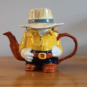 May include: A novelty teapot in the shape of a person, wearing a yellow shirt, dark blue pants, and a tan hat. The teapot has a brown handle and spout, and the person's hands are white. The belt buckle is gold.