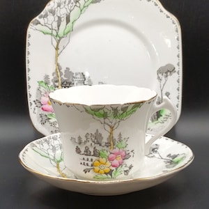 May include: A vintage teacup, saucer, and square plate set. The white porcelain set features a landscape design with trees, a castle, and colorful flowers. The edges are trimmed with gold. The plate measures approximately 8 inches across.