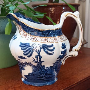 Op de afbeelding: Een blauwe en witte porseleinen roomkan met een gouden rand en een wilgenmotief. De roomkan heeft een handvat en een tuit.