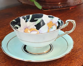 May include: A vintage teacup and saucer set with a floral design. The teacup has a black and white floral pattern with gold accents. The saucer is a light blue color with a gold rim.