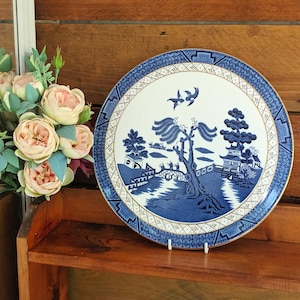 Op de afbeelding: Een blauw en wit decoratief bord met een klassiek Willow-patroon. Het bord heeft een gedetailleerde landschapsweergave met bomen, gebouwen en vogels. Het bord wordt tentoongesteld op een houten plank met een boeket roze en crèmekleurige bloemen.
