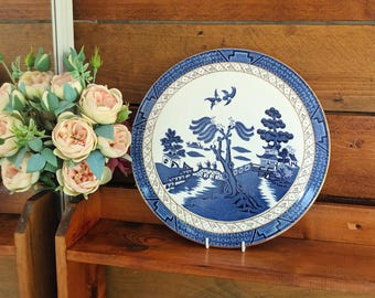 May include: A blue and white decorative plate with a classic Willow pattern design. The plate features a detailed landscape scene with trees, buildings, and birds. The plate is displayed on a wooden shelf with a bouquet of pink and cream-colored flowers.