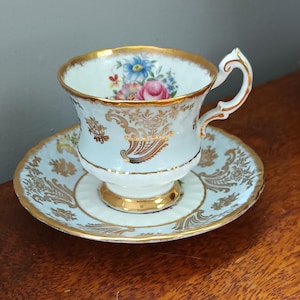 Puede incluir: Un delicado juego de taza y platillo de té con un diseño floral y detalles dorados. La taza y el platillo son blancos con un fondo azul claro.