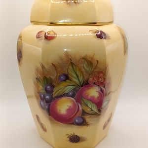 May include: A yellow ceramic jar with a lid, featuring a painted design of fruit and berries. The jar has a octagonal shape and a gold trim around the lid.