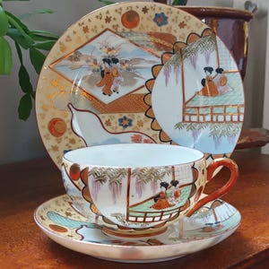Könnte beinhalten: Ein vintage japanisches Tee-Set mit Tasse und Untertasse mit einem zarten Blumen- und Geisha-Design. Tasse und Untertasse sind weiß mit goldenen Akzenten und zeigen eine Szene mit Geishas in einem Garten.