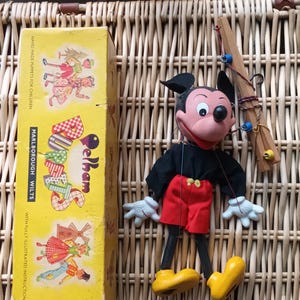 Puede incluir: Una marioneta vintage de Mickey Mouse con una caja amarilla. La marioneta tiene una camisa negra, pantalones cortos rojos, zapatos amarillos y guantes blancos. La caja es amarilla con ilustraciones y el texto "Marlborough Wilts".