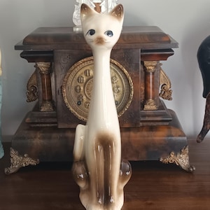 Könnte beinhalten: Eine Vintage-Siamesische-Katzenfigur aus Keramik. Die Katze hat einen cremefarbenen Körper mit braunen Markierungen an Ohren, Gesicht und Pfoten. Sie sitzt mit einem langen, schlanken Hals und leicht geneigtem Kopf. Die Figur steht auf einer Holzoberfläche.