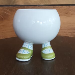 Puede incluir: Una maceta de cerámica blanca con cuerpo redondo y zapatos verdes. La maceta tiene patas con zapatos verdes y blancos y calcetines de lunares azules. La maceta está diseñada para parecerse a una persona.