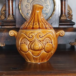 May include: A brown ceramic onion-shaped container with a lid, featuring the word "onions" on the lid. The container has a face-like design with handles on each side. The background includes a vintage clock.