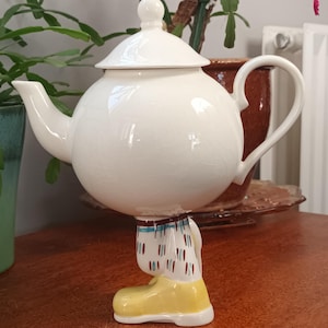 Puede incluir: Una caprichosa tetera de cerámica blanca con cuerpo redondo y tapa. La tetera está sostenida por una base con forma de piernas de personaje de dibujos animados y bota amarilla. La tetera tiene asa y boquilla.