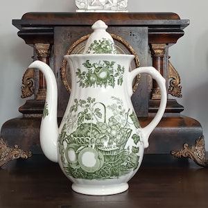 Pode incluir: Bule de cerâmica branco com um padrão verde de frutas e cestas. O bule tem um bico curvo, uma alça e uma tampa com um pequeno botão. O design é clássico, em estilo vintage.