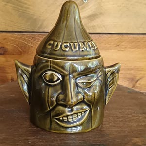 May include: An olive green ceramic jar with a face design, winking eye, and smiling mouth. The lid is inscribed with "CUCUMBER". The jar has ear-like features. The jar is likely for kitchen storage.