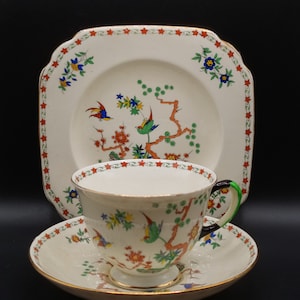 May include: A vintage teacup and saucer set with a floral pattern featuring birds and branches. The set is white with red, green, blue, and yellow accents. The teacup has a black and green handle.
