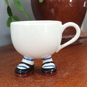 Puede incluir: Una taza de té blanca de cerámica caprichosa con un asa, apoyada sobre dos pies pequeños. Los pies están pintados con zapatos negros, calcetines a rayas azules y blancas y suelas rojas. La taza está sobre una superficie de madera.