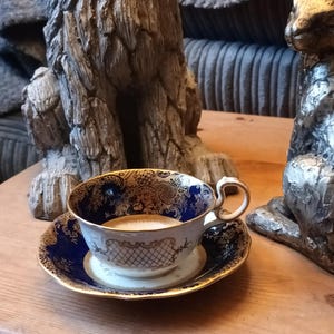 May include: A blue and gold teacup and saucer with a floral design. The cup is filled with a light brown liquid. The teacup and saucer are sitting on a wooden table.