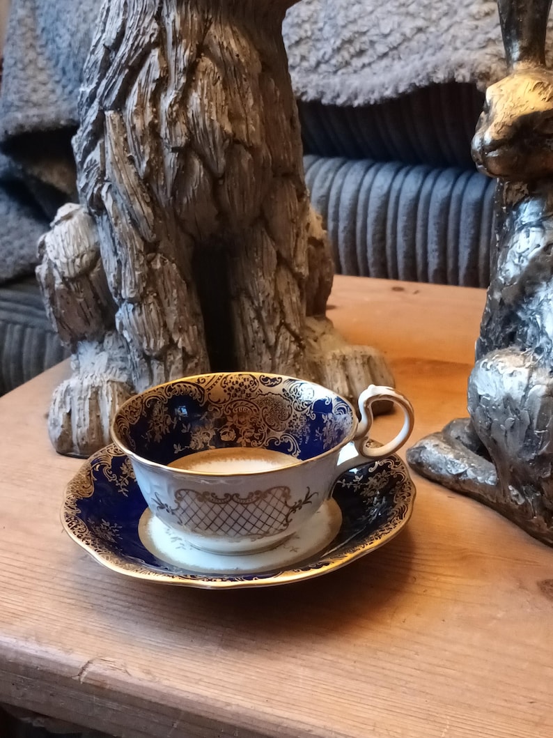 May include: A blue and gold teacup and saucer with a floral design. The cup is filled with a light brown liquid. The teacup and saucer are sitting on a wooden table.