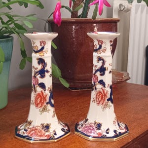 May include: A pair of decorative ceramic candlesticks. The candlesticks are white with floral and blue patterns. The base is hexagonal with a gold trim. The candlesticks are on a wooden surface.