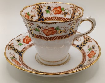 May include: A white teacup and saucer with a floral pattern in shades of orange, blue, and gold. The cup has a scalloped rim and the saucer has a gold rim.