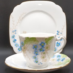 Puede incluir: Un juego de taza y platillo de té blanco con un delicado patrón floral con flores de nomeolvides azules y verdes. El juego tiene un borde dorado.