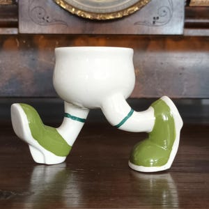 Puede incluir: Un caprichoso huevero de cerámica blanca con piernas y pies verdes y blancos. Las piernas están pintadas con zapatos verdes y rayas verdes en las rodillas. El huevero está sobre una superficie de madera oscura.