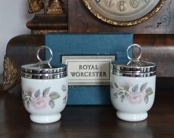 Puede incluir: Dos hueveras Royal Worcester con tapas. Las tazas de porcelana blanca presentan un diseño de rosa rosa y hojas verdes, con adornos plateados y un asa en la tapa. Una caja azul con las palabras "Royal Worcester" está detrás de las tazas.