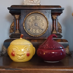 Może przedstawiać: Dwa ceramiczne słoiki do przechowywania żywności. Jeden jest żółty z motywem twarzy i napisem "APPLE SAUCE". Drugi jest bordowy ze słowem "Beets". Oba mają pokrywki.