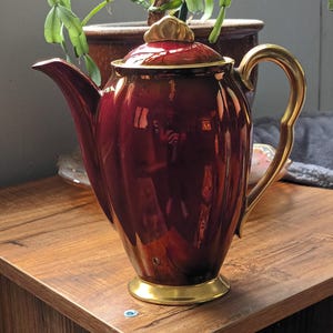 Könnte beinhalten: Eine bordeauxfarbene Keramik-Teekanne mit goldenen Akzenten. Die Teekanne hat einen gebogenen Ausguss, einen goldenen Griff und einen goldenen Sockel. Der Deckel ist ebenfalls bordeauxfarben mit einem goldenen Knauf. Die Teekanne steht auf einer Holzoberfläche.