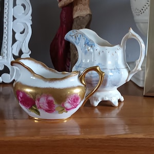 Puede incluir: Dos jarras de crema de porcelana blanca con detalles dorados. Una jarra tiene un diseño floral azul y blanco, la otra tiene un diseño de rosa rosa.
