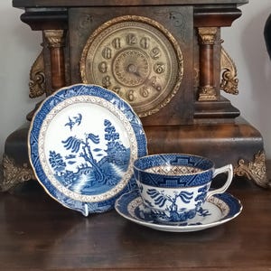Op de afbeelding: Een blauw-wit porseleinen theeservies met een bord, een theekopje en een schotel. De set heeft een gedetailleerd landschapsontwerp met gouden randen. Een antieke houten klok staat op de achtergrond.