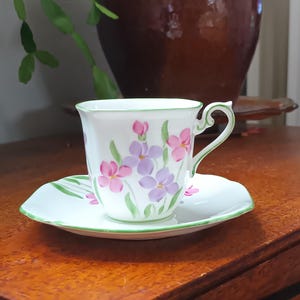 May include: A white teacup and saucer set with a floral design in pink and purple. The cup and saucer have a green trim. The set is placed on a wooden surface.