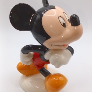 May include: A ceramic figurine of Mickey Mouse, standing with his right foot forward and his left arm bent at the elbow. He is wearing red shorts, yellow shoes, and has a black and white face with large ears.