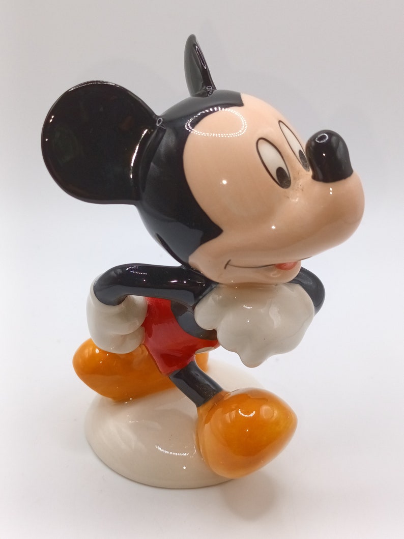 May include: A ceramic figurine of Mickey Mouse, standing with his right foot forward and his left arm bent at the elbow. He is wearing red shorts, yellow shoes, and has a black and white face with large ears.