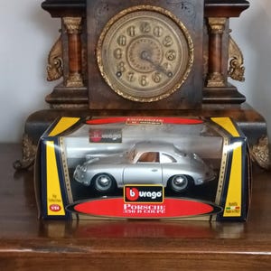May include: A silver Porsche 356 B Coupe model car in a clear display case. The model is 1:24 scale and has the Burago logo. The display case has a black, yellow, and red design. The car is displayed in front of a vintage clock.